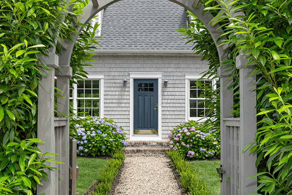 Exterior front view of a home featuring a dark navy blue front entry door with glass panels, surrounded by double-hung windows, an arched wooden trellis, stone pathway, and lush garden landscaping.