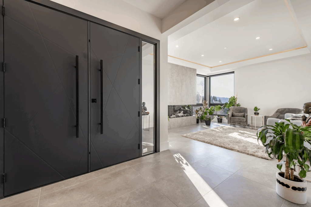 Interior view of a modern home entryway showing a dark charcoal front door with long vertical hardware, adjacent to a bright room with large glass doors, indoor plants, and natural light.