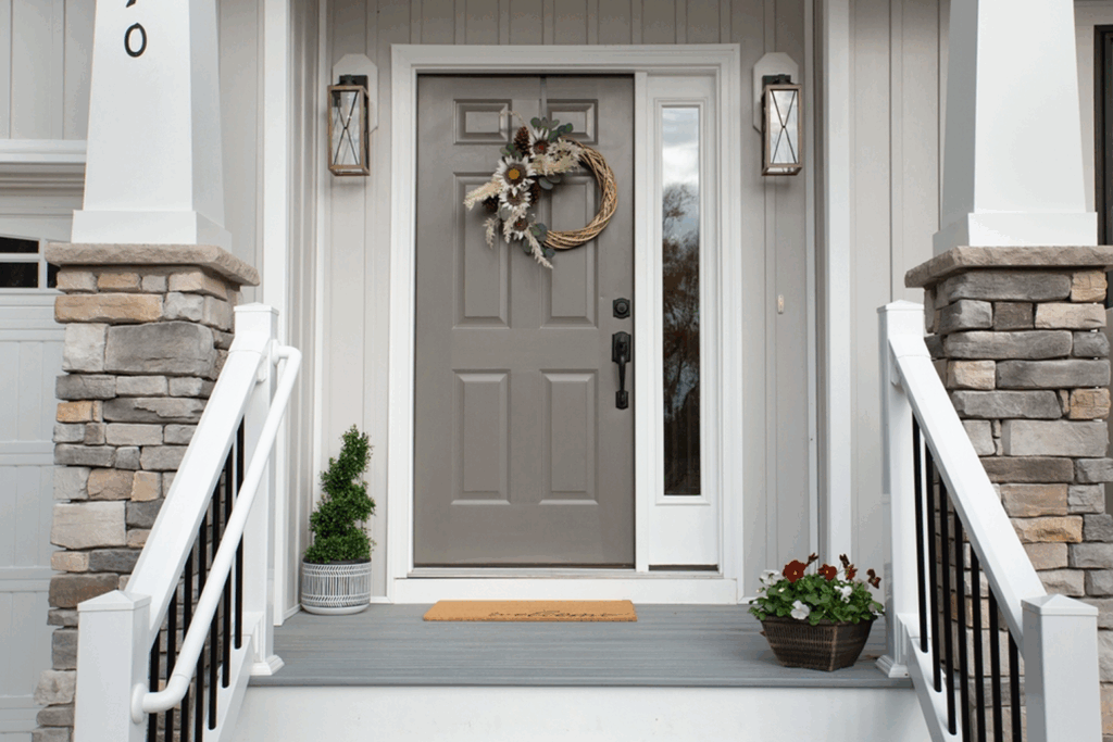 Exterior front porch view featuring a gray front entry door with decorative glass inserts, matching sidelights, stone pillar bases, white railings, and a seasonal door wreath.
