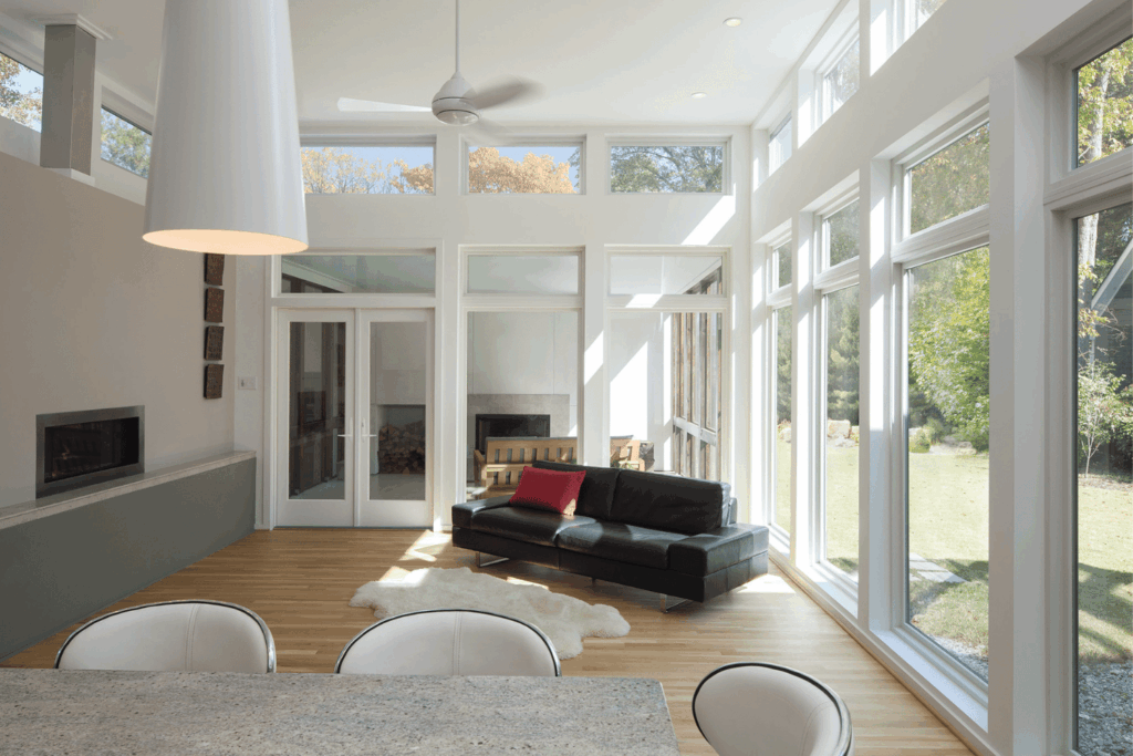 Bright sunroom with expansive floor-to-ceiling windows overlooking a green lawn, dark wood flooring, and a glass-top table with chairs.