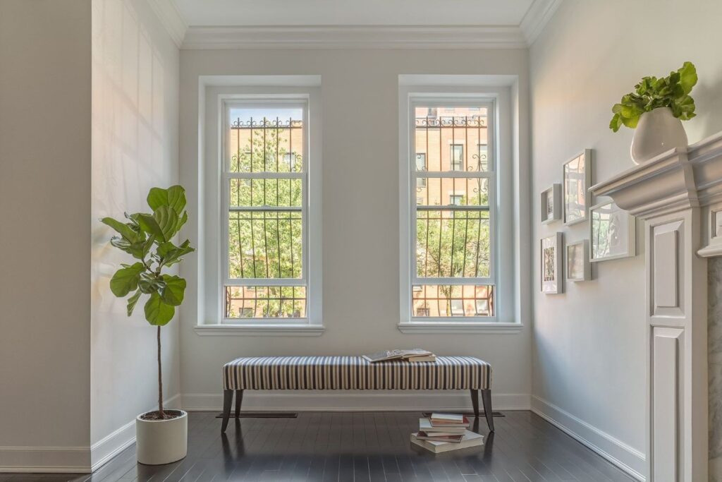 A bright, clean entryway or sunroom space with two double-hung windows. A bench with a striped cushion sits under the windows, flanked by a potted fiddle leaf fig tree on one side and a fireplace mantle with framed photos on the other.