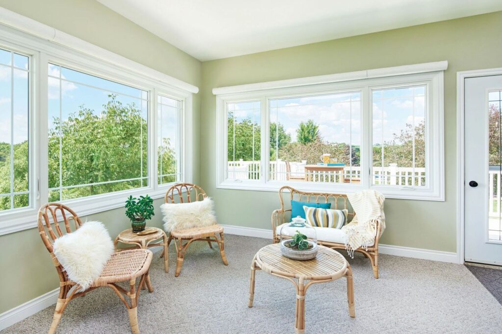 A relaxed sunroom or sitting area with light green walls and large window assemblies on two walls. The room is furnished with two bamboo-style chairs, a loveseat with soft cushions, and a round bamboo coffee table, all creating a comfortable, airy space connected to the outdoors.