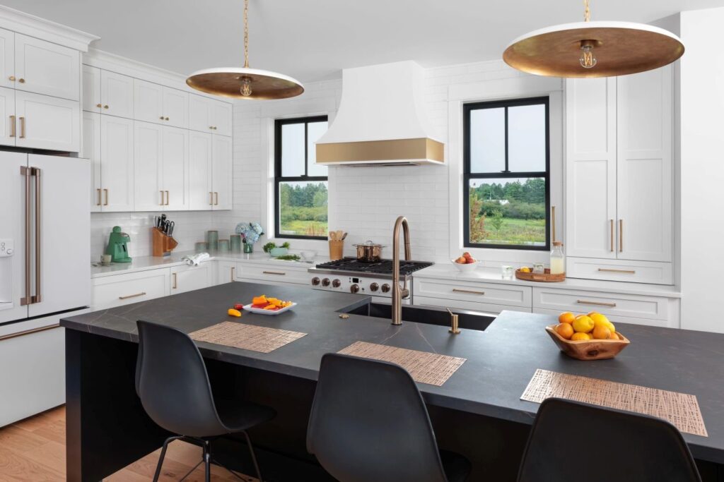 A modern, bright white kitchen with a large dark-colored island, a gas stove, and two large windows above the sink area, allowing plenty of natural light into the space.