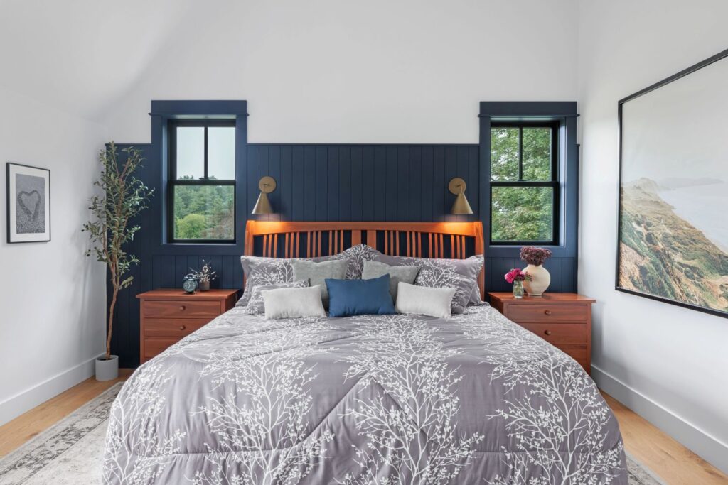 A comfortable, stylish bedroom with dark blue wood-paneled wall accents behind the bed and large windows on either side. The room includes a bed with a grey patterned comforter, two wooden nightstands, and a large piece of wall art depicting a coastal landscape.