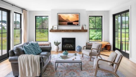 A bright, modern living room featuring large windows with black frames on either side of a white brick fireplace. A cozy sofa and two armchairs are arranged on a patterned rug, creating a warm, inviting space with natural light.