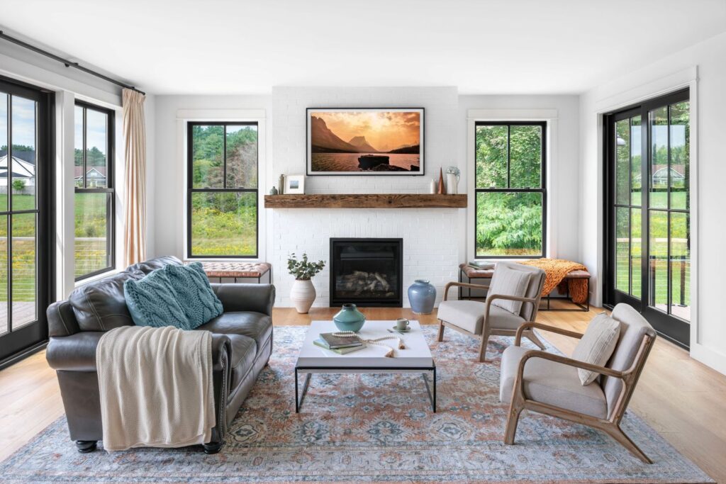 A bright, modern living room featuring large windows with black frames on either side of a white brick fireplace. A cozy sofa and two armchairs are arranged on a patterned rug, creating a warm, inviting space with natural light.